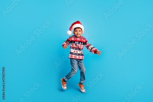 Full size photo of satisfied kid wear red pullover jeans santa hat jumping having fun on christmas party isolated on blue color background