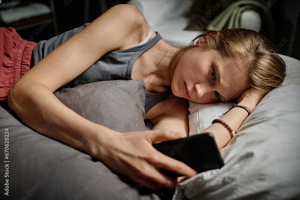 Depressed blond teenage girl looking through cyberbullying messages while lying in bed late at night and suffering from insomnia Depressed blond teenage girl looking through cyberbullying messages while lying in bed late at night and suffering from insomnia