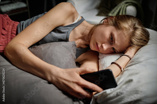 Depressed blond teenage girl looking through cyberbullying messages while lying in bed late at night and suffering from insomnia