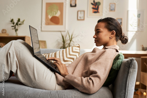 Side view portrait of young Black woman using laptop while lying on couch in cozy home and relaxing