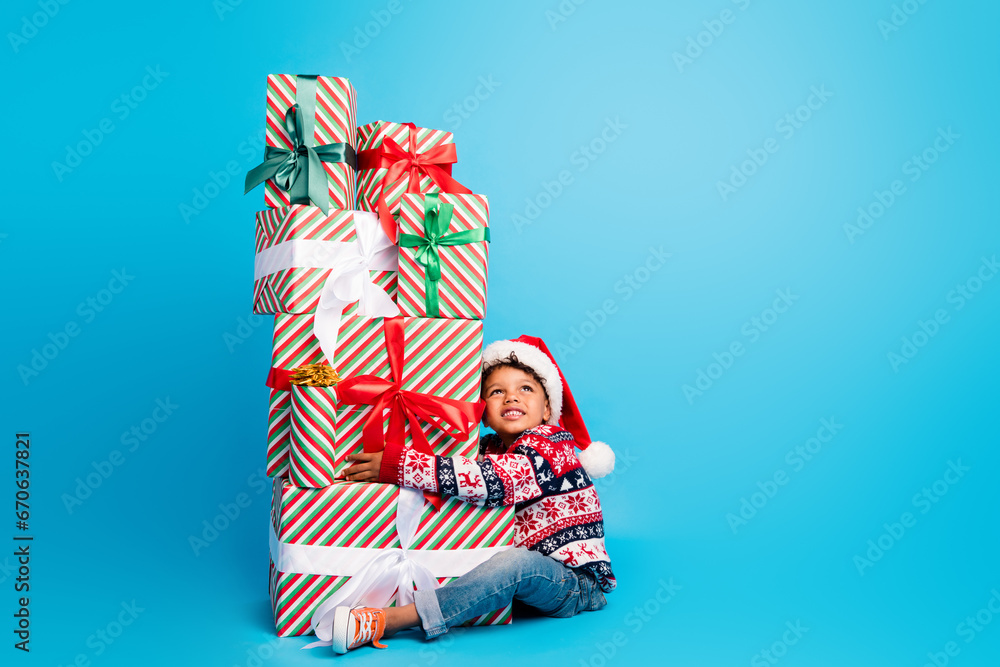 Obraz premium Photo of dreamy small boy dressed ornament sweater santa hat sit cuddle christmas gifts look empty space isolated on blue color background