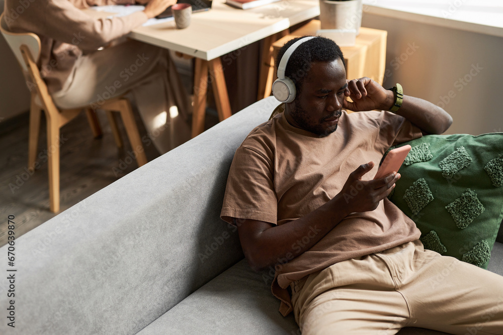 High angle portrait of Black man using smartphone with wireless headphones while listening to music at home, copy space