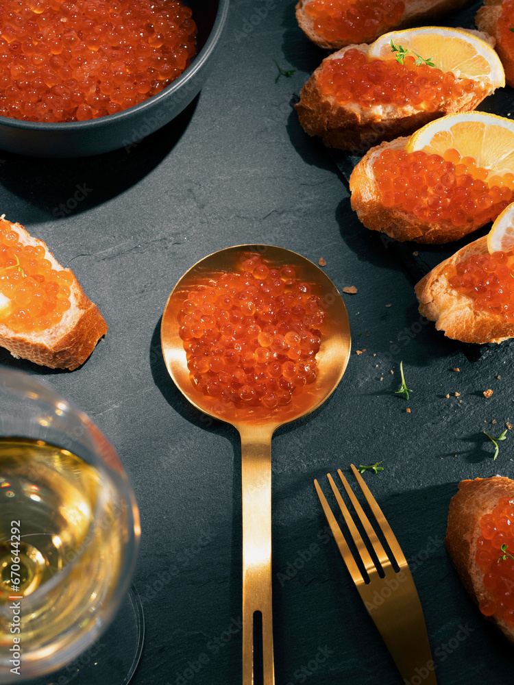 Salmon caviar in a golden spoon and a dark bowl on a dark gray stone background. Red caviar sandwiches with white wine. Gourmet food up close. Seafood and delicatessen.