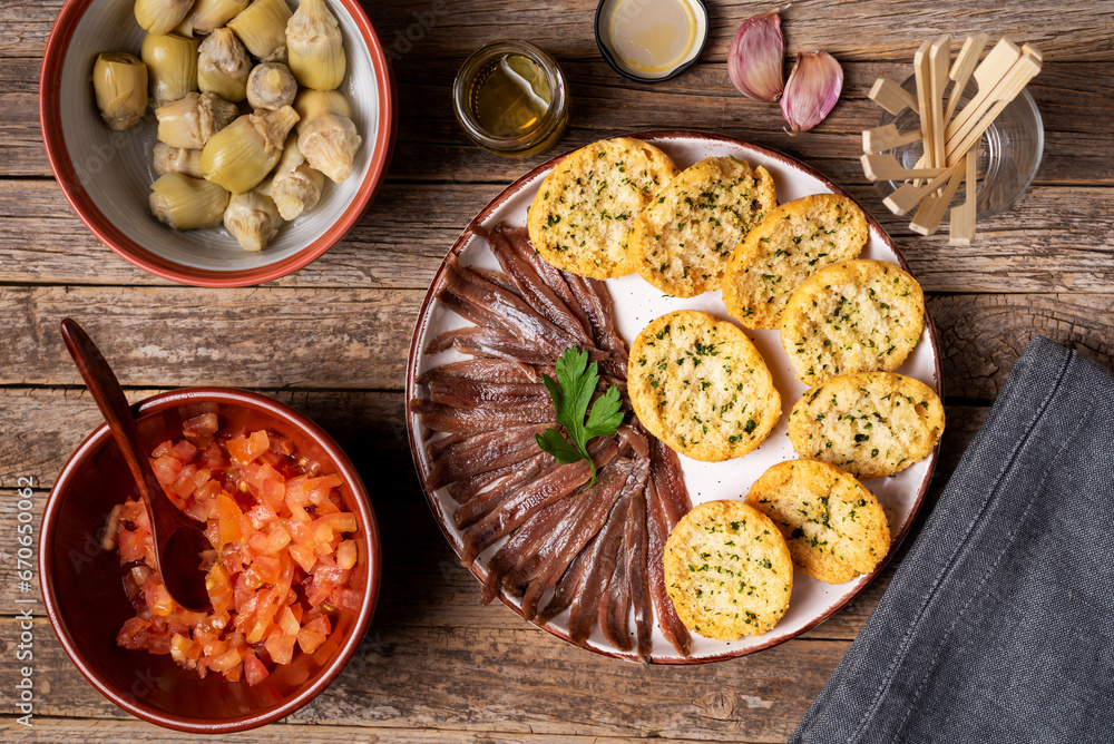 Cantabrian anchovies, toast with parsley, chopped tomato, artichokes, garlic and olive oil, top view, rustic wood background. Cantabrian anchovies, toast with parsley, chopped tomato, artichokes, garlic and olive oil, top view, rustic wood background.