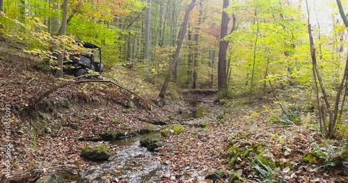All terrain vehicle navigating down a path along a stream in the forest with colorful fall leaves, aerial drone footage