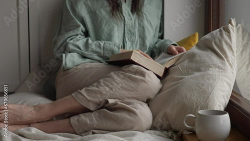 Close up shot woman or girl reading book sitting on floor with pillow near balcony window and drink warm tea or coffee