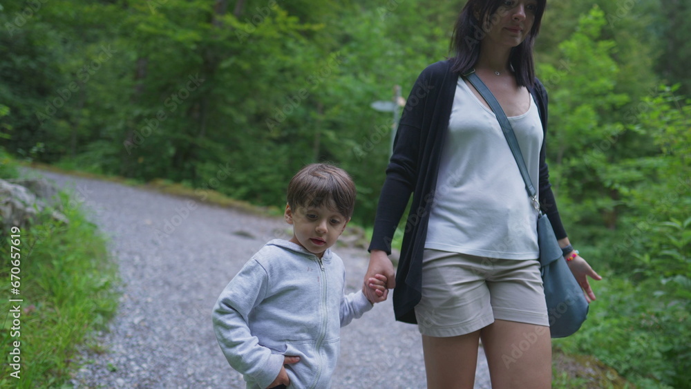 Mother and child holding hands walking in trail path in forest. Mom and son hiking in outdoors, weekend family activity, adventurous journey concept Mother and child holding hands walking in trail path in forest. Mom and son hiking in outdoors, weekend family activity, adventurous journey concept
