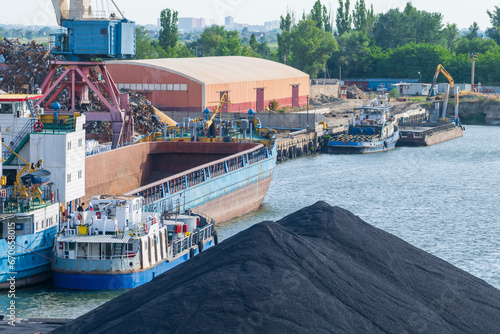 Side view of blue large general cargo ship (bulk carrier) with open deck standing moored in city port in a sunny day. Heap of black coal in the foreground. Soft focus. Freight and shipping theme. photo