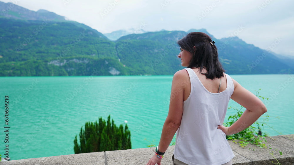 Pensive woman standing by natural lake view with mountains in background. Contemplative female person looking at lakeshire, enjoying nature's calm and serenity
