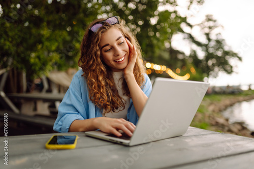 Happy woman in casual clothes working on a laptop in an outdoor cafe near a lake. Young woman freelancer enjoying nature. Tenology concept, working day.