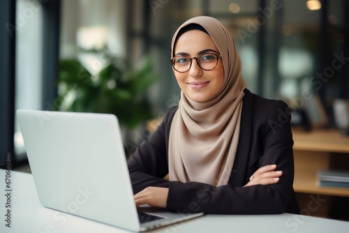 Photo of a young cheerful Muslim woman in eyeglasses wearing hijab working with laptop computer in her modern business office.