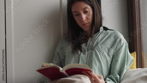 Medium close up shot attractive serious female reading book sitting on the pillow near the window in a rainy day.