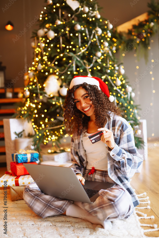 Smiling woman with curly hair with a credit card, gifts and a laptop. A young woman at home near the Christmas tree makes online purchases. Shopping concept, holiday. Smiling woman with curly hair with a credit card, gifts and a laptop. A young woman at home near the Christmas tree makes online purchases. Shopping concept, holiday.