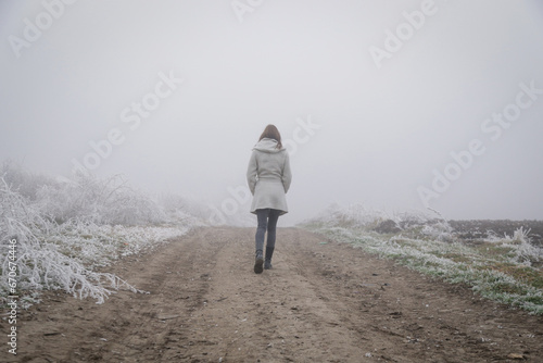 Woman in white coat walking on remote countryside rural road on cold freezing foggy day. Winter weather.