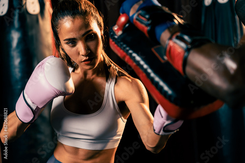 Asian female Muay Thai boxer punching in fierce boxing training session, delivering strike to her sparring trainer wearing punching mitts, showcasing Muay Thai boxing technique and skill. Impetus
