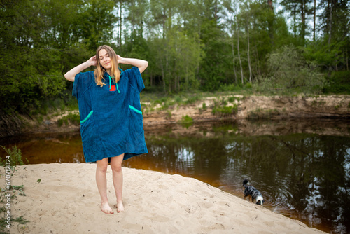 Happy Woman in Blue Poncho Relaxing by Forest River, Curious Dog Exploring the Calm Waters © Uldis Laganovskis