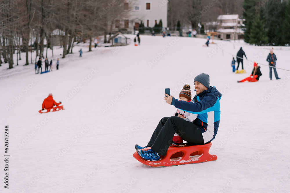Smiling dad taking smartphone selfie of himself and little kid sitting on sled on snowy hill
