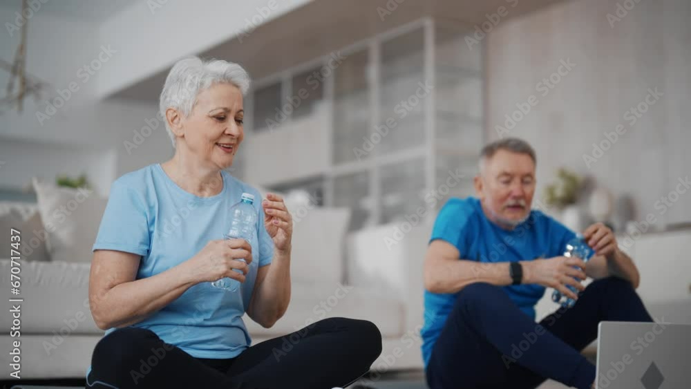 Happy senior couple gray haired man woman talk with fitness trainer in laptop after sport online training sit on mats at home in living room and drinks water. Aged family in sportswear after workout. Happy senior couple gray haired man woman talk with fitness trainer in laptop after sport online training sit on mats at home in living room and drinks water. Aged family in sportswear after workout.