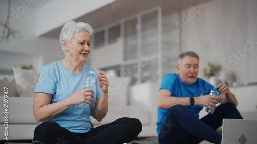 Happy senior couple gray haired man woman talk with fitness trainer in laptop after sport online training sit on mats at home in living room and drinks water. Aged family in sportswear after workout. photo