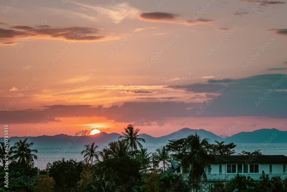 Scenic rose golden glowing Sunset. Half Sun just hid behind mountains. Close up view on palms, sea and mountains at horizon line. Ao Nang, Thailand