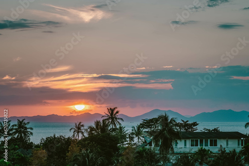 Scenic rose golden glowing Sunset. Half Sun hid in clouds above mountains. Close up view on palms  sea and mountains at horizon line. Ao Nang  Thailand