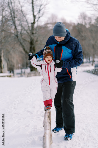 Little girl walks along a narrow log in the park  holding on to her father hands in a snowy park