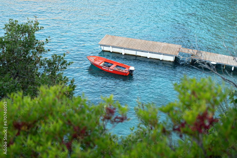 View to calm sea bay from above through some coniferous trees, red rescue boat docked near small wooden pier