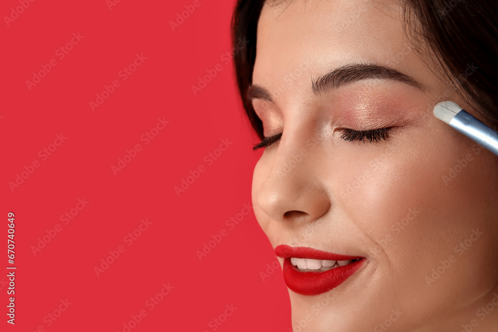 Beautiful young woman with brush applying eyeshadows on red background