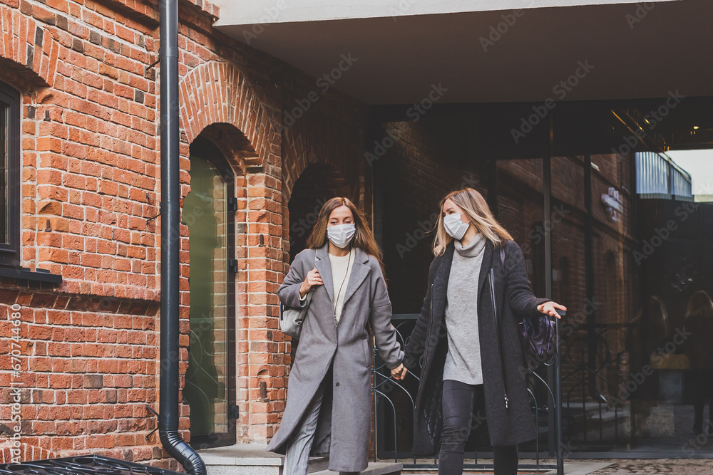 Naklejka premium Two young stylish women in medical masks talking and laughing while walking the street in town. Friends strolling outdoor in city