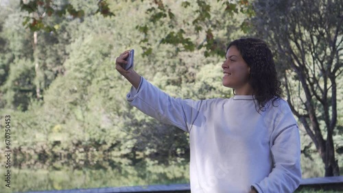 Young Caucasian brunette girl in white sweatshirt taking a smiling selfie with her cell phone in a natural setting