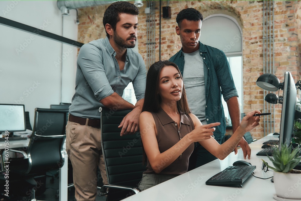Teamwork. Young asian beautiful woman pointing at computer monitor and discussing reports with her coworkers while sitting in modern office Teamwork. Young asian beautiful woman pointing at computer monitor and discussing reports with her coworkers while sitting in modern office