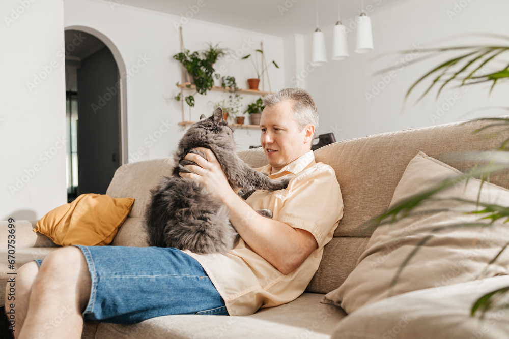 A middle-aged man sits and cuddles with a gray fluffy cat on the sofa in the living room and relaxes after work. Adored pets A middle-aged man sits and cuddles with a gray fluffy cat on the sofa in the living room and relaxes after work. Adored pets