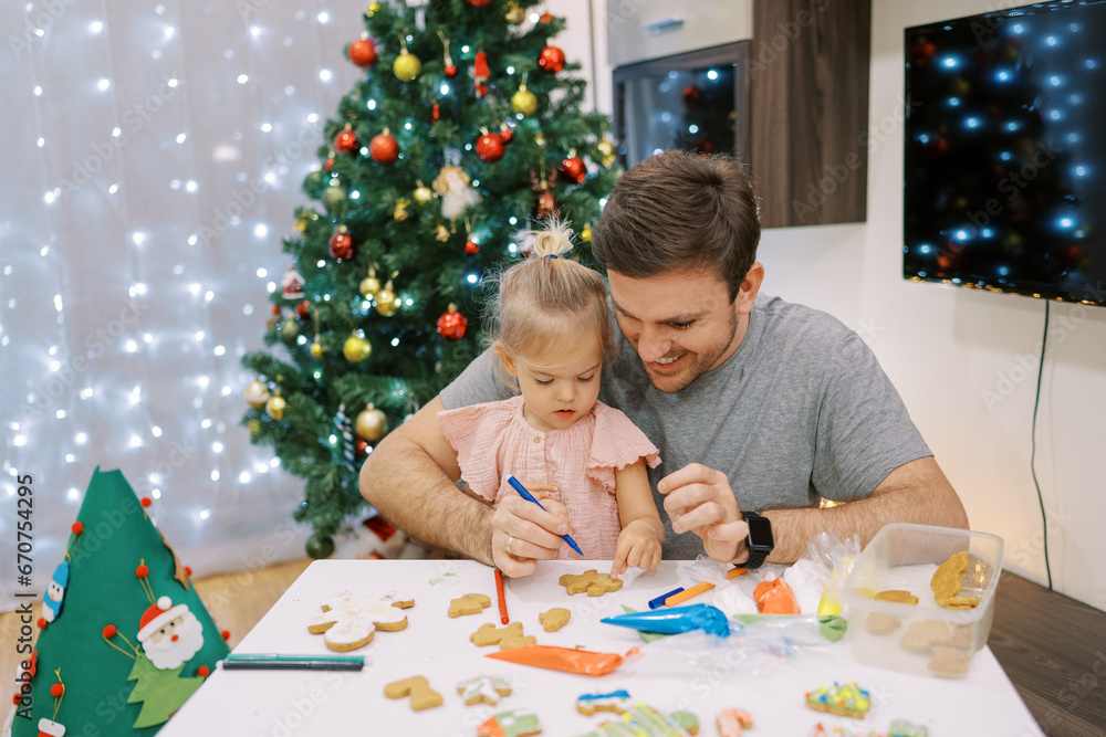 Little girl sits on the lap of her dad drawing with a felt-tip pen on a Christmas cookie at the table