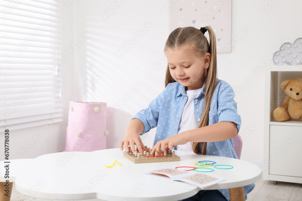 Motor skills development. Smiling girl playing with geoboard and rubber bands at white table in room. Space for text Motor skills development. Smiling girl playing with geoboard and rubber bands at white table in room. Space for text