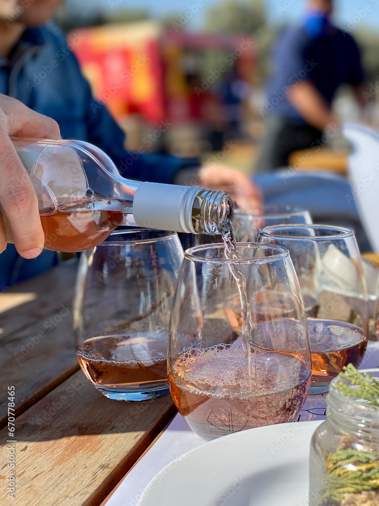 Rosé wine being poured into a glass. The bottle is held by a person's hand and the glass is one of several on an outdoor table.