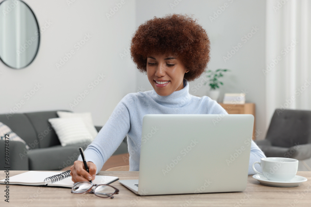 Beautiful young woman using laptop and writing in notebook at wooden desk in room Beautiful young woman using laptop and writing in notebook at wooden desk in room