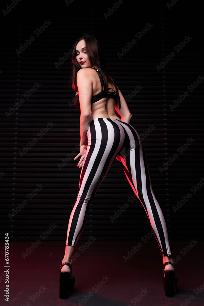 Beautiful girl in a black bra, leather belts and leggings in black and white lines poses bending over against the background of a wooden wall. Back view. Party concept, night club Beautiful girl in a black bra, leather belts and leggings in black and white lines poses bending over against the background of a wooden wall. Back view. Party concept, night club