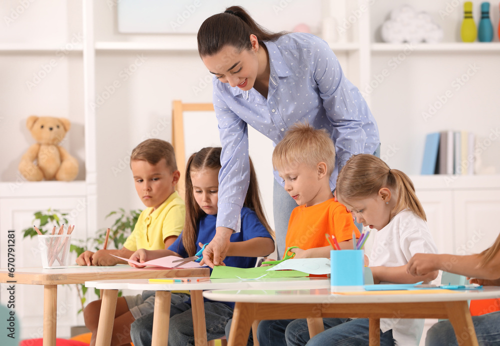 Nursery teacher with group of cute little children drawing and cutting paper at desks in kindergarten. Playtime activities Nursery teacher with group of cute little children drawing and cutting paper at desks in kindergarten. Playtime activities