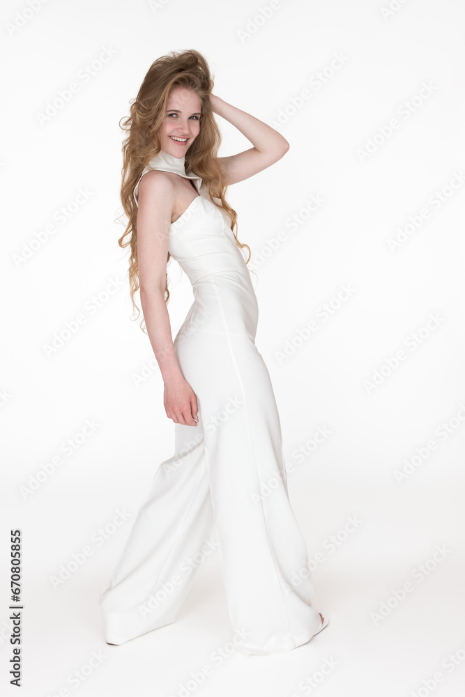 Playful and smiling young fashion model dressed in white long jumpsuit with hand behind head looking over shoulder at camera. Happiness woman with long blonde hair. Studio shot on white background Playful and smiling young fashion model dressed in white long jumpsuit with hand behind head looking over shoulder at camera. Happiness woman with long blonde hair. Studio shot on white background