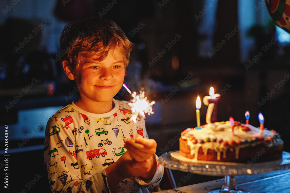 Adorable happy blond little kid boy celebrating his birthday. Child blowing seven candles on homemade baked cake, indoor. Birthday party for school children, family celebration of 7 years