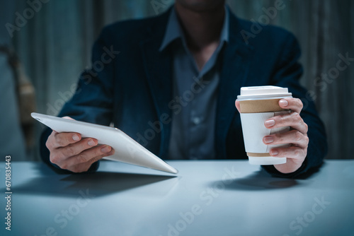 Businessman working in corporate workplace having reading data document on ipad screen while holding drinking coffee cup sitting in low light dark indoor office as young adult executive photo