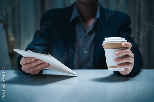 Businessman working in corporate workplace having reading data document on ipad screen while holding drinking coffee cup sitting in low light dark indoor office as young adult executive