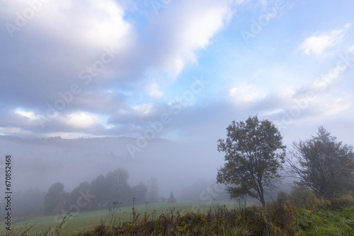 Beautiful autumn scenic panorama of a foggy valley in the Carpathian mountains in the early morning. Grass hill with trees in the foreground.