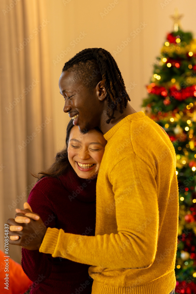Happy, romantic diverse couple dancing at home and smiling by christmas tree