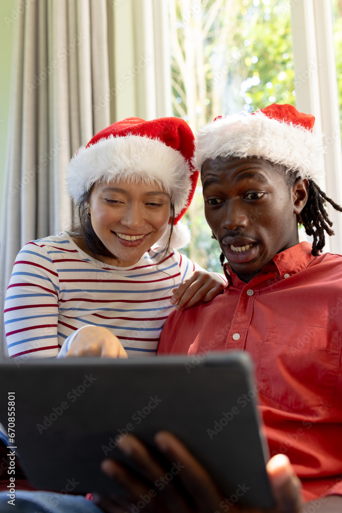 Happy, surprised diverse couple in christmas hats having tablet video call in sunny living room Happy, surprised diverse couple in christmas hats having tablet video call in sunny living room