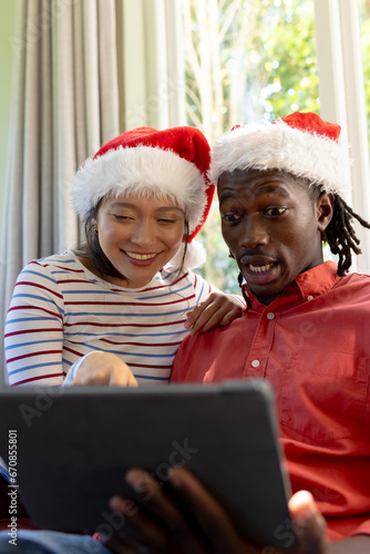 Happy, surprised diverse couple in christmas hats having tablet video call in sunny living room