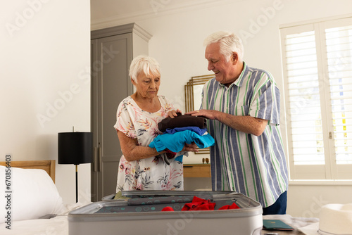 Happy caucasian senior couple packing suitcase in sunny bedroom at home