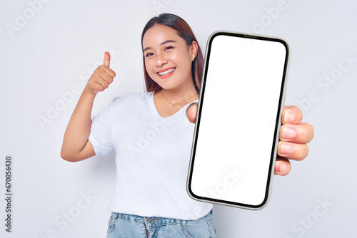 Beautiful cheerful Asian girl wearing casual white t-shirt showing mobile phone with blank black screen and raising thumb up isolated on white background. People lifestyle concept