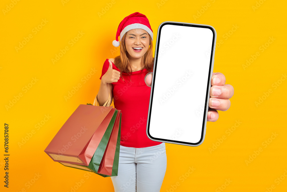 Φωτογραφία Smiling young Asian woman in Santa Claus hat holding blank screen phone and shop