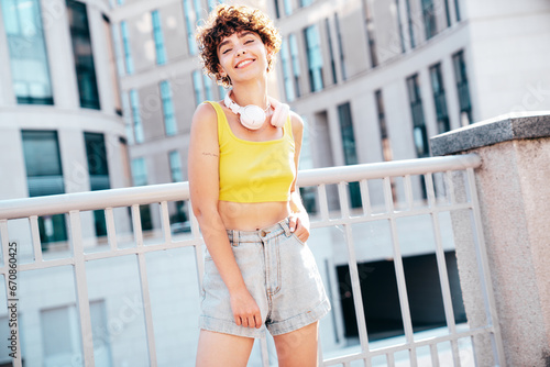 Young beautiful smiling hipster woman in trendy summer clothes. Carefree woman with curls hairstyle  posing in the street at sunny day. Positive model outdoors. Listens music at her headphones on neck